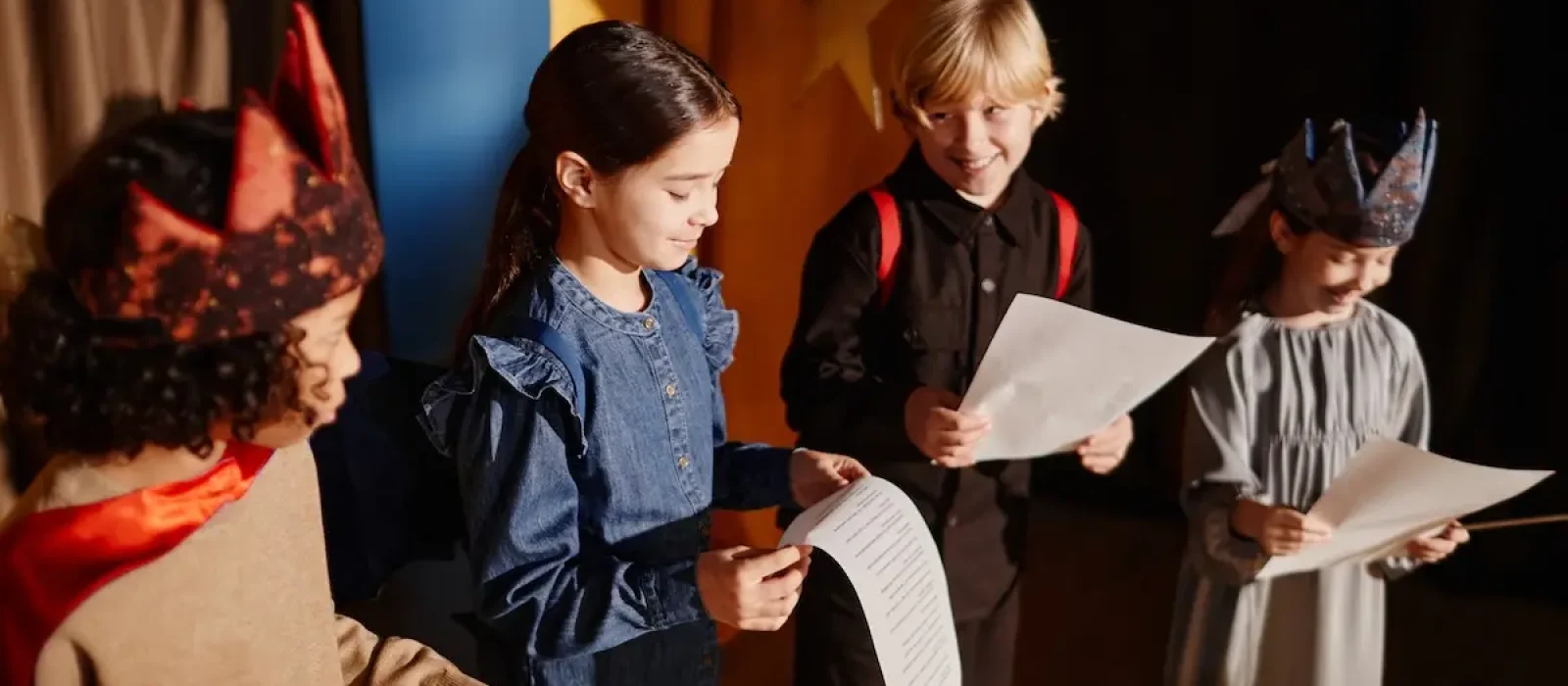 Children Participating in Classroom Theater Activity Young students rehearsing for school play, some wearing costumes and holding scripts, happily engaging in theater activity. Wooden floor and colorful backdrop create vibrant atmosphere
