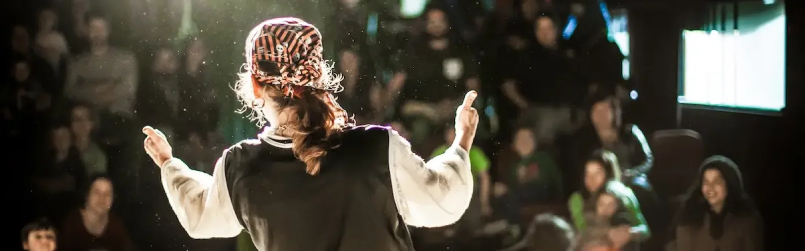 A performing arts entertainer is seen from the back in selective focus, dressed as a pirate on stage during a comedy act with blurry audience at back.